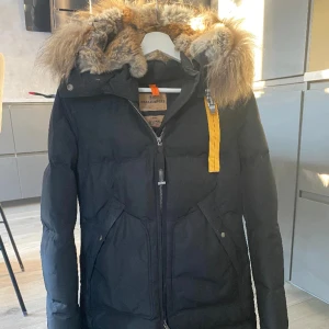 Svart dunjacka Parajumpers med päls - Svart dunjacka från Parajumpers med fluffig päls runt huvan och en gul detalj vid kragen. Jackan har dragkedja framtill, stora fickor och en klassisk vinterlook. Perfekt för kalla dagar och riktigt snygg till jeans eller boots.