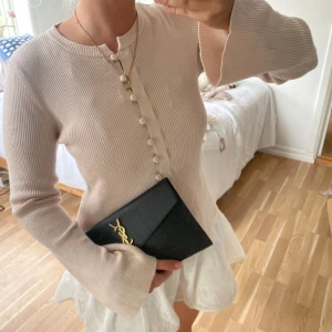 Topp 🤍🤍🍨 - Stilren svart kuvertväska från YSL med klassisk guldlogga framtill. Väskan har en elegant, enkel design i slätt material och stängs med ett lock. Perfekt att bära i handen för en chic look.