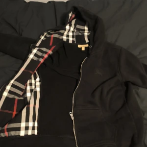 Svart hoodiejacka från Burberry - Svart hoodiejacka från Burberry med klassiskt rutigt foder i huvan. Jackan har dragkedja framtill, fickor och diskret broderad logga på bröstet. Tillverkad i 100% bomull för en skön och avslappnad känsla. Perfekt för dig som gillar stilren streetwear.