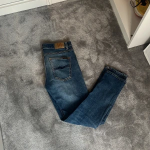 Blå jeans från Nudie Jeans - Säljer ett par klassiska blå jeans från Nudie Jeans med broderade bakfickor👖 Modellen är lean dean vilket är straight/slim. Jeansen har snygg blå wash. Jeansen har ett par före detta hål som blivit proffesionellt lagade på nudie jeans i stockholm👍