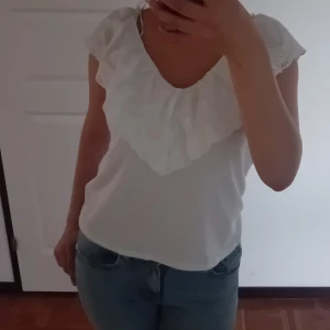 Vit blus med volang från H&M - Supersöt vit blus från H&M med bred volang och broderade detaljer längs ringningen. Blusen har korta ärmar med spetskant och en luftig passform som känns perfekt för varma dagar. Stilren och romantisk look som lyfter vilken outfit som helst. Diskuterar gärna pris😊