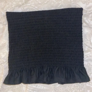 Svart bandeau tubtopp med volang - Supersöt svart bandeau tubtopp med smockad struktur och volangkant nedtill. Perfekt att styla med höga jeans eller kjol för en trendig look. Toppens stretchiga material gör den bekväm och enkel att bära.