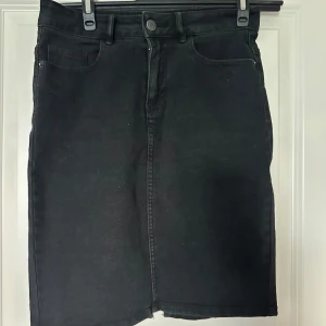 Svart jeanskjol från Vero Moda - Svart jeanskjol från Vero Moda i klassisk modell med fem fickor, bälteshällor och dragkedja fram. Kjolen har en liten slits bak och är i stretchigt denimtyg som sitter snyggt. Perfekt till sneakers eller boots.