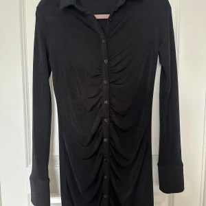 Svart skjortklänning från Gina Tricot - Svart skjortklänning från Gina Tricot med långa ärmar och knappar hela vägen framtill. Klänningen har snygg rynkdetalj längs knappraden och klassisk krage. Mjuk och stretchig känsla, perfekt för en trendig look.