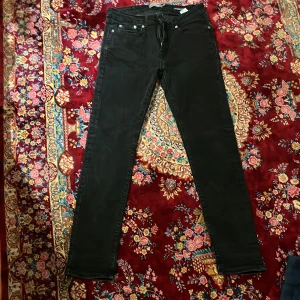 Svarta slim fit jeans Dressmann - Svarta jeans från Dressmann i slim fit-modell. Klassisk femficksdesign med dragkedja och knapp framtill. Jeansen har raka ben och är tillverkade i ett stretchigt bomullsmaterial för en bekväm passform. Perfekta för en clean och stilren look. För barn. Storlek 29-30. Använda ca 3 ggr.