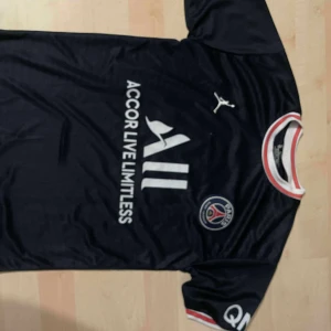 PSG Messi #30 matchtröja Jordan S - Säljer en mörkblå Paris Saint-Germain fotbollströja med Messi och nummer 30 på ryggen. Tröjan har vita och röda detaljer vid krage och ärmslut, PSG-logga på bröstet och Jordan-logga. Tillverkad i lätt och ventilerande material, perfekt för fotbollsälskare.