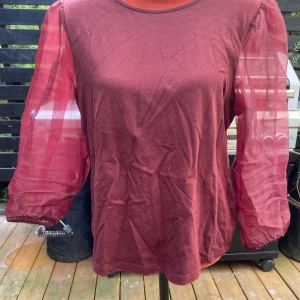 Vinröd topp med puffärmar från KappAhl - Vinröd blus med långa, genomskinliga ärmar. Blusen har en rund halsringning och en avslappnad passform. Längd: 61 cm ﻿Bystvidd: 48 cm.  Inga bilder när den sitter på mig! Katter finns i hemmet!