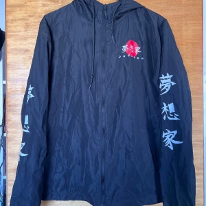 Svart vindjacka - Svart vindjacka med huva och dragkedja. Japan inspirerat tryck och stor röd cirkel med texten 'Dreamer' på bröstet, ryggen och ärmarna. Jackan är i lätt polyester och har en loose passform, perfekt för dig som gillar streetstyle.