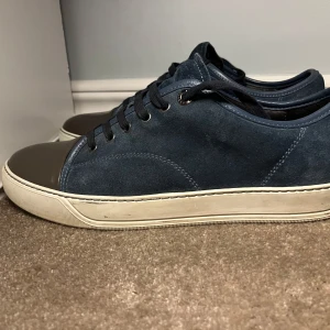 Lanvin skor - Snygga sneakers i mörkblå mocka med grå, glansig tå och vita sulor. Skorna har svarta snören och en klassisk låg profil. Perfekta för dig som gillar stilrena detaljer och en modern look. Jag har använt de få gånger och tycker att den lila slitning som finns gör de väldigt snyggt. Inga defekter skick 9/10. 