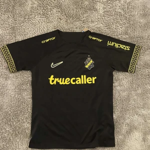 AIK svart matchtröja Nike M - AIK:s officiella svarta matchtröja från Nike med gula detaljer och tryck. Truecaller och SVEA-loggor, klubbmärke på bröstet och jubileumstryck 1934-2024 i nacken. Korta ärmar med gul mönsterkant. Tillverkad i lätt och ventilerande polyester.