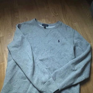Grå sweatshirt från Polo Ralph Lauren - Klassisk grå sweatshirt från Polo Ralph Lauren med rund halsringning och broderad marinblå logga på bröstet. Tröjan har långa ärmar och ribbade muddar vid ärmslut och nederkant. Perfekt basic att ha i garderoben. Använd varsamt, och inköpt inte alls längesedan