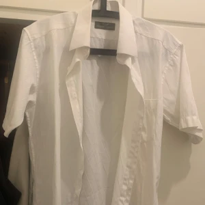 Vit kortärmad skjorta från Giorgio Armani - Vit kortärmad skjorta från Giorgio Armani i 100% bomull. Skjortan har diskreta diagonala ränder, klassisk krage, bröstficka med broderad logga och knäppning framtill. Perfekt för dig som vill ha en clean och stilren look.