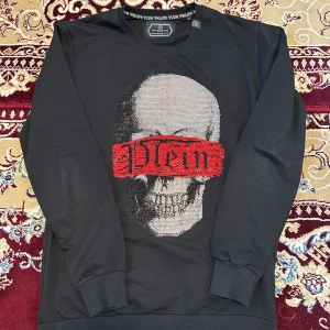 Svart Philipp Plein tröja med döskalle - Svart långärmad tröja från Philipp Plein med Platinum Cut. Framsidan har en stor döskalle med glittrande detaljer och röd text över ögonen. Tröjan har ribbade muddar och logotypdetaljer i nacken och på sidan. Perfekt för dig som gillar edgy streetstyle.