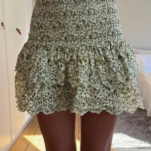 Vit och grön blommig volangkjol från Zara  - Supersöt kort kjol med volanger och broderade blommor i grönt på vit botten. Kjolen har smock i midjan och flera lager med vågig spetskant. Perfekt för en somrig look och lätt att matcha med en enkel topp.