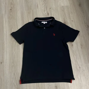Marinblå pikétröja från U.S. Polo Assn. - Snygg marinblå pikétröja från U.S. Polo Assn. med klassisk krage, två knappar och röd broderad logga på bröstet. Tröjan har diskreta röda och vita detaljer på insidan av kragen och vid slitsarna nertill. Perfekt för en stilren och sportig look.