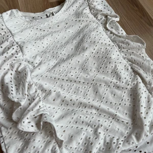 Vit blus med volang från Pieces - Supersöt vit blus från Pieces med broderade hålmönster och volangdetaljer längs axlarna. Ärmlös modell med rund halsringning och luftig känsla, perfekt för varma dagar. Ger en fräsch och trendig look till din outfit.