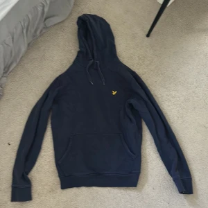 Marinblå hoodie från Lyle & Scott - Snygg marinblå hoodie från Lyle & Scott med klassisk gul logga på bröstet. Perfekt för dig som gillar en enkel och stilren tröja.