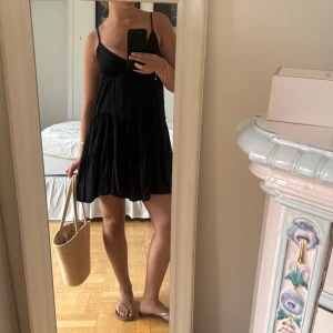 Svart kort sommarklänning med tunna band - Supersöt svart kort klänning med tunna axelband och v-ringning. Klänningen har en luftig passform och volangdetaljer i kjolen, perfekt för varma dagar. Enkel och stilren design som är lätt att matcha med accessoarer.