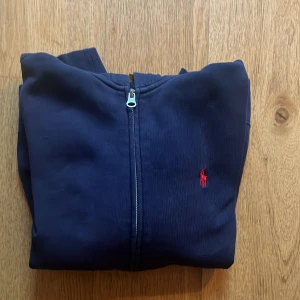 Marinblå hoodie från Polo Ralph Lauren - Snygg marinblå hoodie från Polo Ralph Lauren med dragkedja och klassisk röd logga på bröstet. Tröjan har huva, känguruficka och långa ärmar. Perfekt för chill dagar och enkel att matcha med jeans eller joggers. Hoddien är använd mycket men fortfarande i bra kvalitet. Storlek M (10-12)