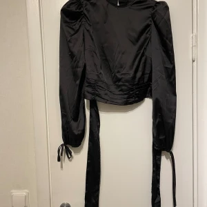 Svart omlottopp från SHEIN med knyt - Snygg svart omlottopp från SHEIN i glansig polyester. Toppen har långa puffärmar med knyt vid handlederna och långa band i midjan som knyts i ryggen. Croppad modell med coola detaljer och stilren look. Perfekt för dig som vill sticka ut lite extra.