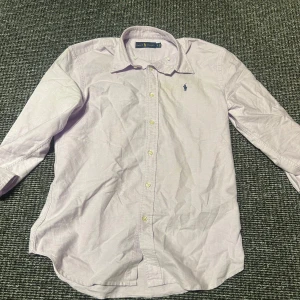 Ljuslila skjorta från Ralph Lauren  - Säljer en klassisk ljuslila skjorta från Ralph Lauren med lång ärm och knappar framtill. Skjortan har en snygg krage och den ikoniska broderade loggan på bröstet. Tillverkad i mjuk bomull och passar perfekt.
