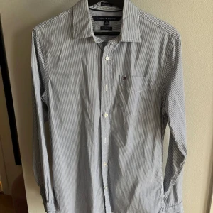Randig skjorta från Tommy Hilfiger - Snygg skjorta från Tommy Hilfiger med smala blå och vita ränder. Klassisk krage, långärmad och med knappar framtill. Bröstficka med liten logga. Skjortan är i bomull och har en normal passform, perfekt för en stilren look.