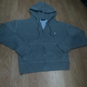 Grå Ralph lauren zip hoodie - En grå Ralph lauren zip hoodie. Bra kvalitet knappt använt. Tröjan har broderad logga på bröstet, två fickor framtill och mjukt foder på insidan. Perfekt för chill dagar och enkel att matcha med jeans eller joggers. Storlek L men skulle passar bra om man har storlek M också.