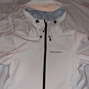 Vit vindjacka Peak Performance - Snygg vit vindjacka från Peak Performance med svart dragkedja och justerbar huva. Jackan har logga på bröstet och ärmen, samt praktiska detaljer som dragsko vid huvan. Perfekt för blåsiga dagar och stilren look. Några små fläckar vid magen som går bort i tvätt