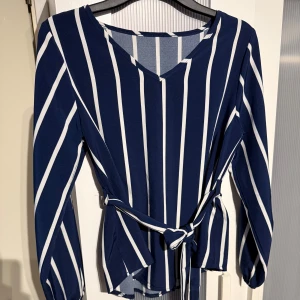 Marinblå randig blus med knytband - Snygg marinblå blus med vita vertikala ränder och v-ringning. Långa ärmar med resår vid handlederna och ett knytband i midjan som ger en schysst siluett. Perfekt för dig som gillar stilrena och trendiga plagg.
