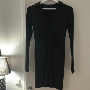 Svart långärmad klänning med knappar - Svart långärmad klänning från Nelly, med snygg rynkning framtill och knappar längs hela framsidan. Klänningen är v-ringad, med ett stretchigt material. Har även en liten slits vid händerna. Använd endast 1 gång, priset kan diskuteras!