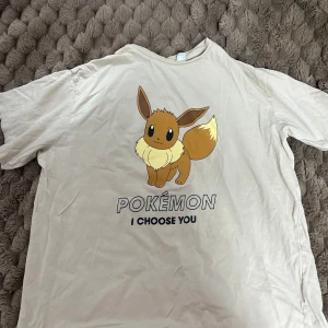 Beige Pokémon Eevee t-shirt H&M - Beige t-shirt från H&M med stor Eevee-tryck och texten 'POKÉMON I CHOOSE YOU' på framsidan. Klassisk passform med rund hals och korta ärmar. Perfekt för dig som älskar Pokémon och vill ha en chill och snygg look.