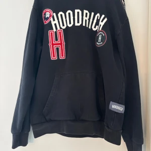 Svart hoodie från Hoodrich med patchar - Svart hoodie från Hoodrich med stora broderade patchar och tryck i vitt och rött framtill. Klassisk känguruficka, huva och ribbade muddar. Hoodrich-logga på ärmen och coola detaljer som ger streetkänsla. Perfekt för dig som gillar urban stil.