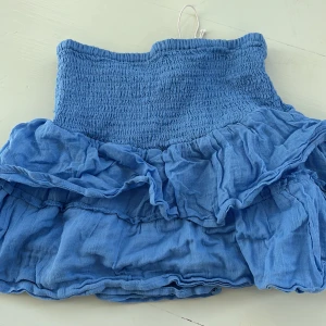 Blå volangkjol från Gina Tricot - Söt blå kjol från Gina Tricot med volangdetaljer och elastisk midja. Perfekt för en avslappnad och stilren look. Kjolen har en kort längd och är tillverkad i ett lätt och luftigt material.