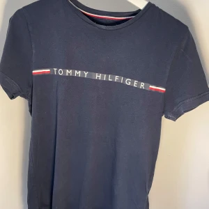 Mörkblå t-shirt från Tommy Hilfiger - Snygg mörkblå t-shirt från Tommy Hilfiger med klassisk logga och ränder i rött och vitt på bröstet. Perfekt för en stilren och avslappnad look. T-shirten har korta ärmar och rund halsringning.