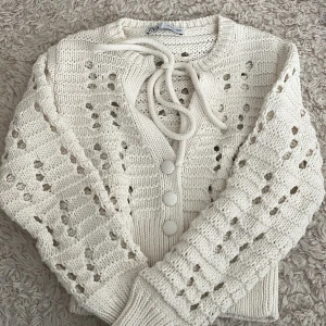 Vit stickad kofta från Zara - Säljer en söt vit stickad kofta från Zara med knytning framtill och dekorativa knappar. Koftan har ett fint hålmönster och långa ärmar med ribbade muddar. 🤍