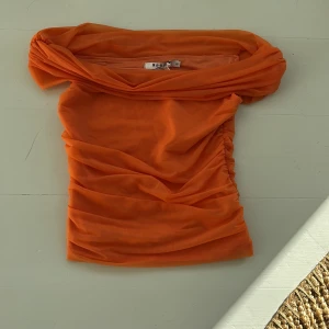 Orange offshoulder topp från NA-KD - Snygg orange offshoulder topp från NA-KD med rynkade detaljer på sidorna. Perfekt för att sticka ut med en härlig färg och stil. Tillverkad i ett lätt och luftigt material.