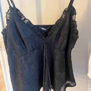 Svart blommig linne från Gina Tricot - Snygg svart linne från Gina Tricot med blommigt mönster och volangdetaljer. Toppen har smala axelband och en v-ringad design som ger en elegant look. Perfekt för en kväll ute eller en speciell tillställning.