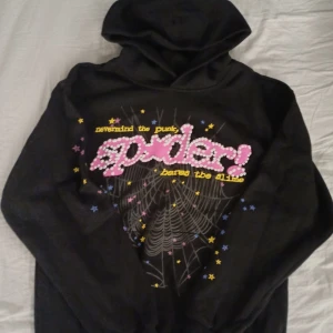 Svart spider hoodie - En svart spider hoodie med vit spindelnät och rosa text. Runt om finns det gula, blåa och rosa stjärnor. Klistermärken sitter kvar, vilket bevisar att den är knappast använd. Axel bredden är 47 cm och hoodien är 67 cm lång.                      Priset kan diskuteras. Hoodien är använd en gång och det var bara för att testa den. Perfekt för sommaren, våren och hösten. Skriv för mer bilder!!!