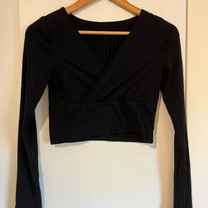 Svart v-ringad ribbad topp - Snygg svart ribbad omlottopp med långa ärmar. V-ringad och är perfekt för en stilren look. Toppen har en omlottdesign som ger en elegant touch.