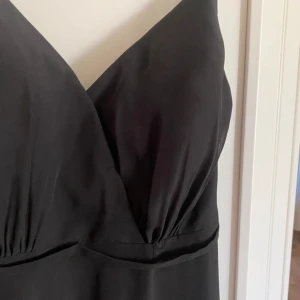 Svart långklänning med v-ringning - Elegant svart långklänning med djup v-ringning och smala axelband. Klänningen har en flytande passform och är perfekt för formella tillfällen.