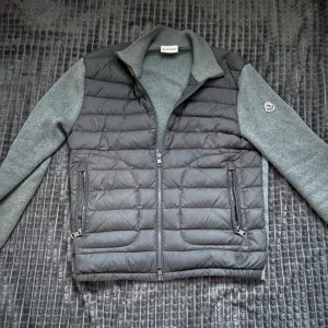 Grå cardigan från Moncler - Snygg grå cardigan från Moncler med dragkedja och quiltad design. Västen har gråa ärmar och en broderad logga på ärmen. Perfekt för kyligare dagar och till våren som kommer nu kan gå ner lite i pris annars hör av er vid minsta fundering!