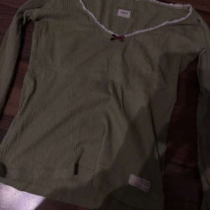 Grön ribbad topp från Odd Molly - Säljer en grön ribbad topp från Odd Molly med v-ringning och en söt rosett framtill. Toppen har långa ärmar och en dekorativ spetskant vid halsen. 