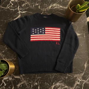 Mörkblå stickad tröja från Polo Ralph Lauren - Säljer en snygg mörkblå stickad tröja från Polo Ralph Lauren med en stor amerikansk flagga på framsidan och RL-broderi. Tröjan har långa ärmar och ribbade muddar vid ärmslut och nederkant. Perfekt för en stilren look! Den är väldigt kort ochså så det passar korta personer