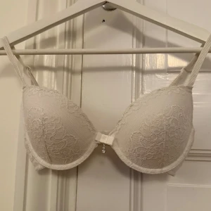 Vit spets-bh med detalj - Elegant vit bh med spetsdetaljer och en söt detalj framtill. Justerbara axelband och hakknäppning bak för perfekt passform. Perfekt för en romantisk look.