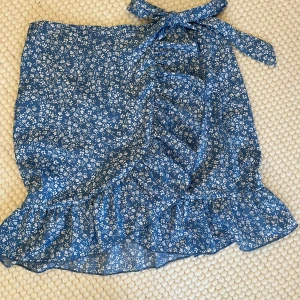 Blå blommig omlottkjol - blå omlottkjol med vitt blommönster och volangdetaljer. Kjolen har en knytning i midjan och en dragkedja baktill