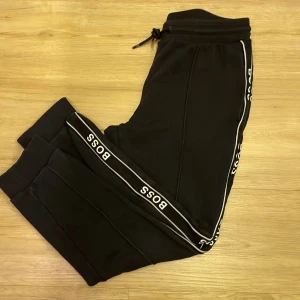 Svarta mjukisbyxor från Boss - Snygga svarta mjukisbyxor från Boss med vita detaljer längs sidorna. Byxorna har en elastisk midja med snörning och ribbade muddar vid bensluten. Perfekta för en avslappnad stil.