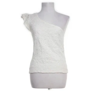 Vit spetsprydd oneshoulder topp - Snygg vit oneshoulder topp med spetsdetaljer. Perfekt för en stilren look med en unik touch. Den har en asymmetrisk design som ger en modern känsla.