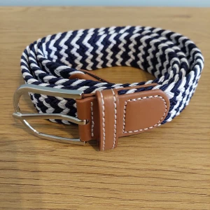 Randigt flätat bälte med läderdetaljer - Snyggt flätat bälte i blått och vitt med bruna läderdetaljer. Perfekt för att ge en stilren touch till din outfit. Bältet har ett klassiskt spänne i silverfärg och är justerbart för bästa passform.