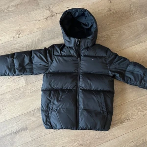 Svart pufferjacka från Tommy Hilfiger - Säljer en svart pufferjacka från Tommy Hilfiger med huva och dragkedja. Jackan har en klassisk design med märkets logga på bröstet och insidan. Väldigt fint skick!  