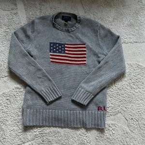 Grå stickad tröja från Polo Ralph Lauren - Säljer en stilren grå stickad tröja från Polo Ralph Lauren med en amerikansk flagga på bröstet. Tröjan har långa ärmar och ribbade muddar vid ärmslut och nederkant. Perfekt för en avslappnad och trendig look.
